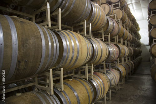 Horizontal Close Up Front three-Quarter View, 4-High Stacked Rows of French Oak Wine Barrels, Cases of Wine in Background, Clean Commercial Lighted Warehouse, Cement Floor