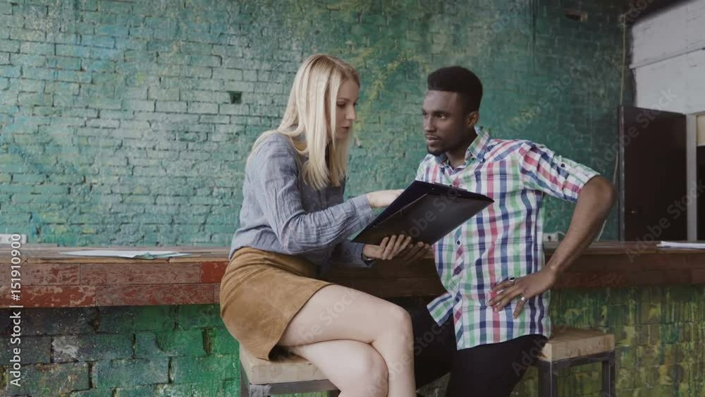 Young african man and caucasian woman sits near the bar counter at modern office and discussing the documents. Young african man and caucasian woman sits near the bar counter at modern office and discussing the documents.