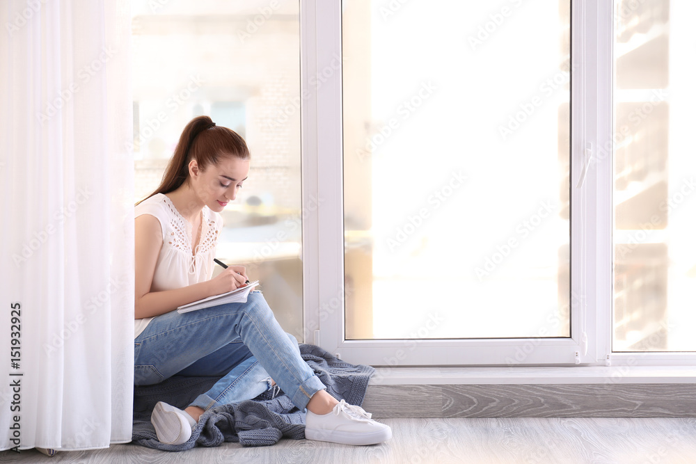 Beautiful young woman with pen and notebook sitting on windowsill Beautiful young woman with pen and notebook sitting on windowsill