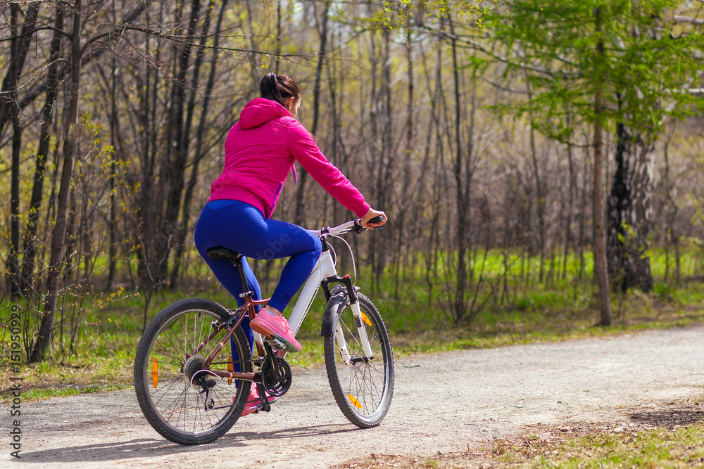 Sporty young woman in a bright pink jacket and jeans rides a bike through the forest on a summer day. Back view