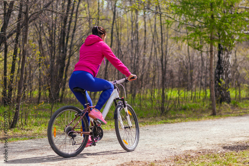 Sporty young woman in a bright pink jacket and jeans rides a bike through the forest on a summer day. Back view