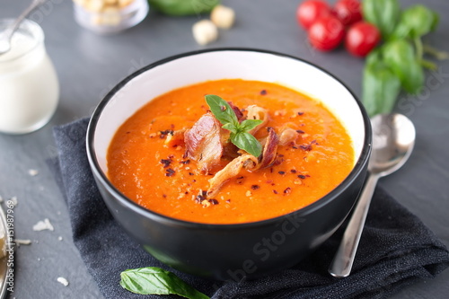 Thick tomato soup with basil and fried bacon in a black ceramic bowl on a grey abstract background. Healthy eating concept Fresh tomato soup with creme fraiche