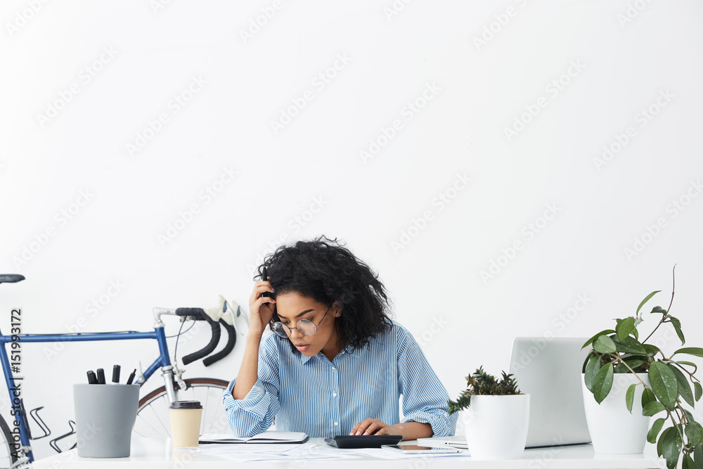 © wayhome.studio - Serious dark-skinned woman accountant wearing formal shirt and eyeglasses calculating bills, sitting at white desk and looking at sheets of paper with concentrated expression, analysing figures © wayhome.studio - Serious dark-skinned woman accountant wearing formal shirt and eyeglasses calculating bills, sitting at white desk and looking at sheets of paper with concentrated expression, analysing figures