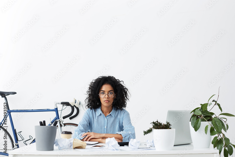 Serious mixed race columnist with Afro haircut suffering from writer's block, sitting in office with crumpled paper balls scattered all over white desk, looking at camera with frustrated expression Serious mixed race columnist with Afro haircut suffering from writer's block, sitting in office with crumpled paper balls scattered all over white desk, looking at camera with frustrated expression