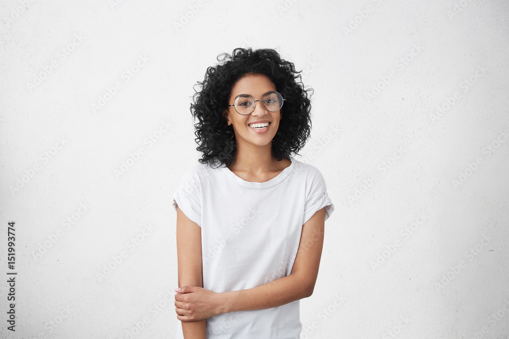 People, lifestyle, youth and happiness concept. Shy pretty girl with shaggy black hair and dark skin wearing stylish eyeglasses, feeling happy after boy who she likes invited her out on date People, lifestyle, youth and happiness concept. Shy pretty girl with shaggy black hair and dark skin wearing stylish eyeglasses, feeling happy after boy who she likes invited her out on date