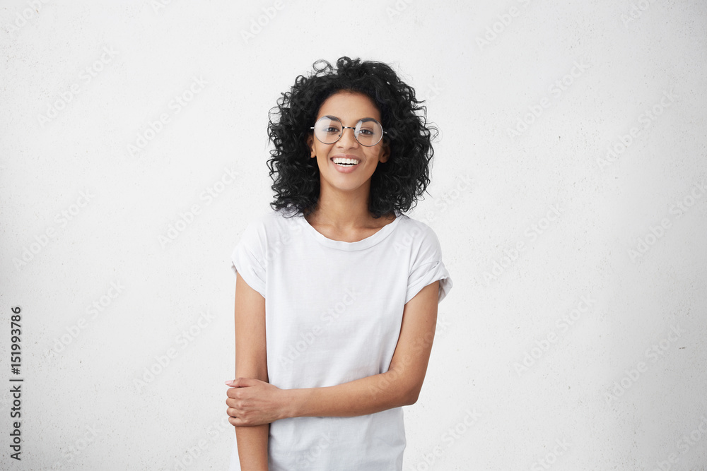 © wayhome.studio  - Indoor portrait of charming young mixed race female model dressed casually feeling shy and awkward during her first photo shoot, standing at white blank studio wall with copy space for content
