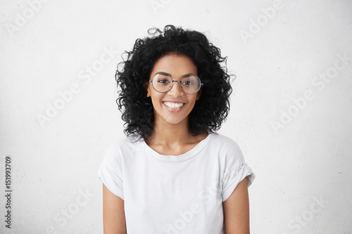 Indoor portrait of beautiful brunette young dark-skinned woman with shaggy hairstyle smiling cheerfully, showing her white teeth to camera while feeling happy and carefree on her first day-off photo