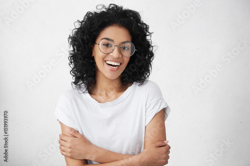 Cheerful young good looking woman with clean dark skin and black shaggy hair posing indoors with crossed arms, smiling broadly with her white straight teeth, laughing at good joke, wearing casual top