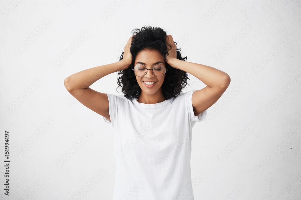 Happy beautiful young woman with dark skin touching her black hair and keeping eyes closed while feeling joyful and excited about something good happened, can't believe her ears, dressed casually Happy beautiful young woman with dark skin touching her black hair and keeping eyes closed while feeling joyful and excited about something good happened, can't believe her ears, dressed casually