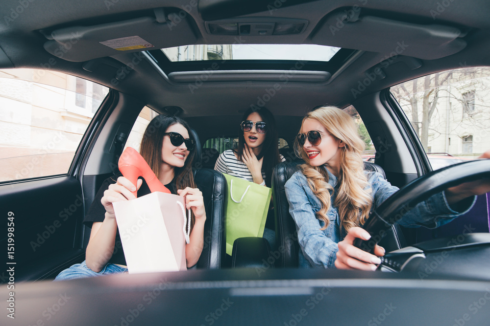 Three beautiful young cheerful women holding shopping bags and looking at each other with smile while sitting in car