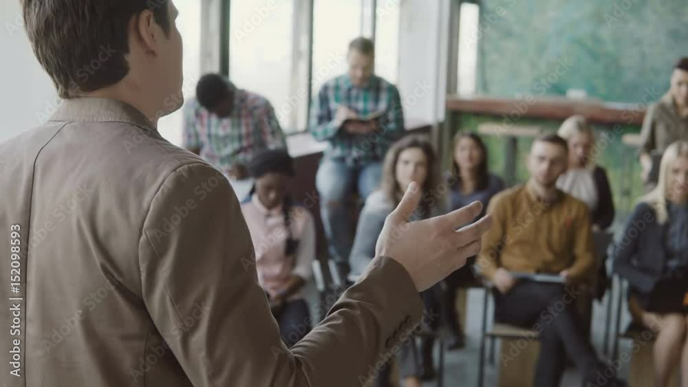Close-up view of young businessman speaking at seminar at modern office. Mixed race group of people listening the male. Close-up view of young businessman speaking at seminar at modern office. Mixed race group of people listening the male.