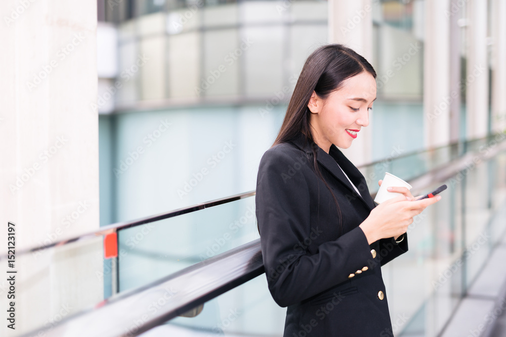 Young Asian woman with smartphone standing against street blurred building background. Fashion business photo of beautiful girl in black casual suite with phone and cup of coffee and use smartphone Young Asian woman with smartphone standing against street blurred building background. Fashion business photo of beautiful girl in black casual suite with phone and cup of coffee and use smartphone