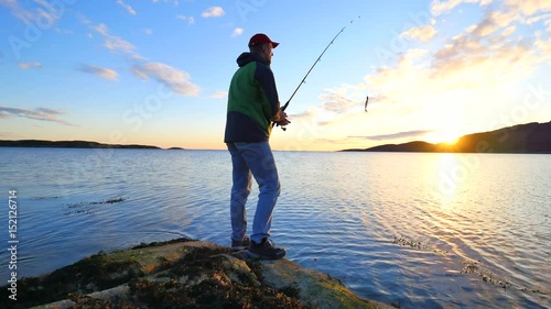 The active man is fishing on sea from the rocky coast. Fisherman check pushing bait on the fishing line, prepare rod and than throw lure into peacefull water. Fisherman silhouette at sunset