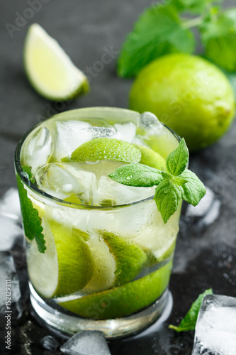Cold refreshing summer drink with lime, mint and ice cubes in a glass on a dark stone background. Mojito. Lemonade. Detox. Mineral water. Selective focus