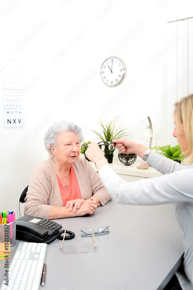 Mature female doctor ophthalmologist in her office trying eyeglasses to a patient elderly senior woman Mature female doctor ophthalmologist in her office trying eyeglasses to a patient elderly senior woman