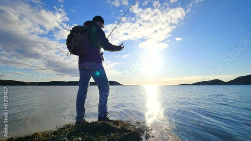 The fisherman with green sporty backpack is checking fishing equipment.  Man prepare pushing bait on fishing line and throw lure far into calm sea water. Fisherman enjoy afternoon sport activities.