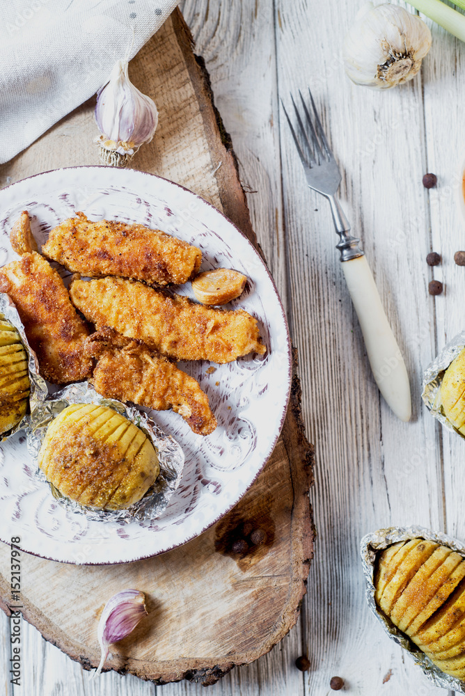 Hearty and delicious dinner, a baked potato with chicken sticks in crumbs, sauce, garlic and spices on a light wooden background Hearty and delicious dinner, a baked potato with chicken sticks in crumbs, sauce, garlic and spices on a light wooden background