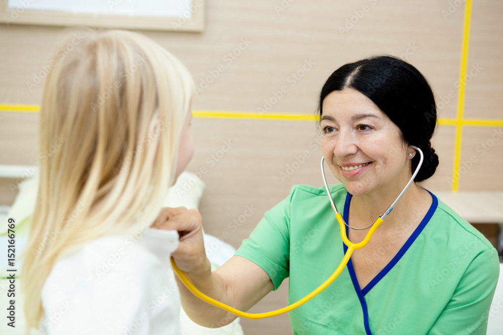 Mature female pediatrician examining little girl with a stethoscope at the clinic Mature female pediatrician examining little girl with a stethoscope at the clinic