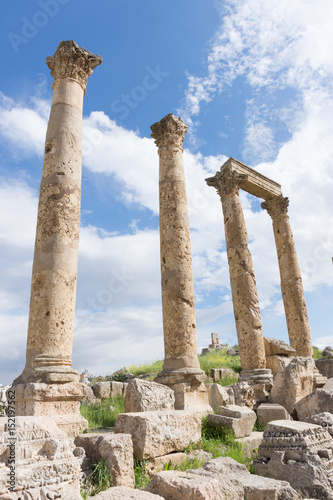 Ancient Roman columns with capitals and a stone beam amidst a pile of rubble in Jerash Jordan. Blue sky with clouds is above. 