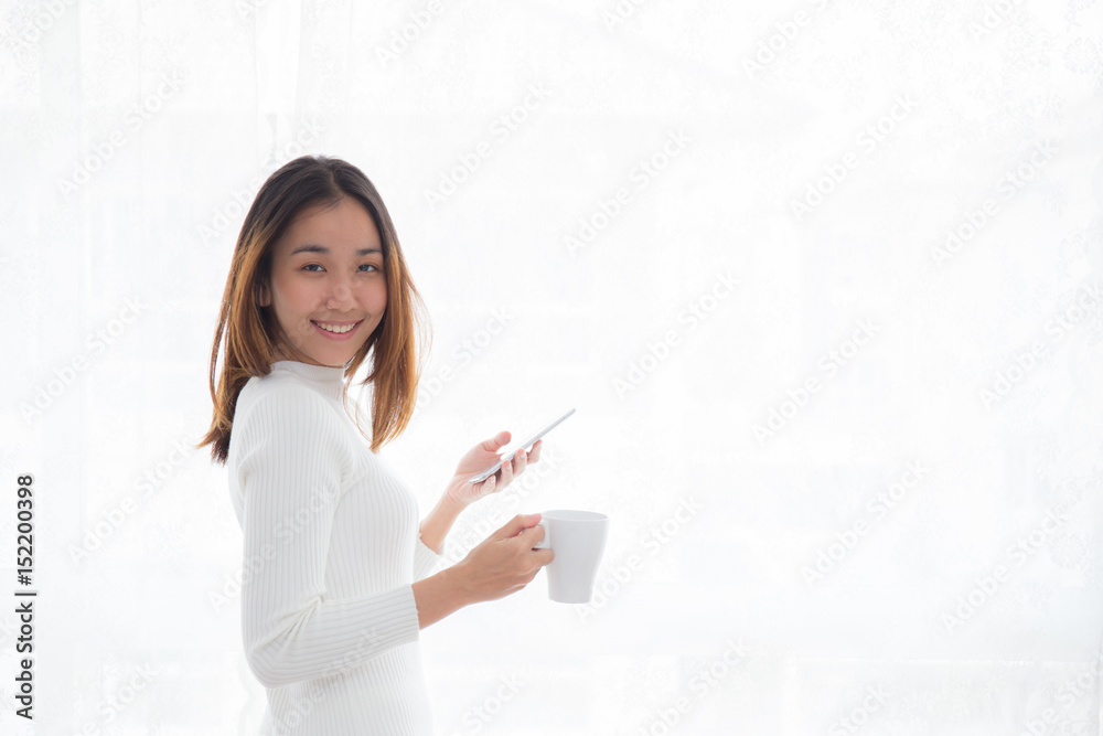 Beautiful young woman in white sweater drinking coffee and holding smartphone at a window. Beautiful young woman in white sweater drinking coffee and holding smartphone at a window.
