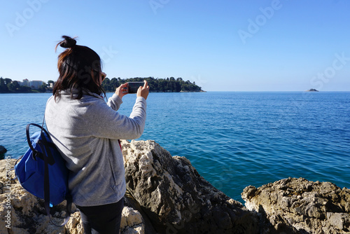 Asian woman standing on the rock taking a photo of adriatic sea with smart phone