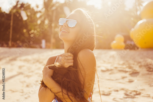 close-up portrait of a beautiful young brunette in sunglasses girl on a sunny day in the summer on the beach by the sea smiling at sunset in denim shorts and top with print US flag lifestyle 