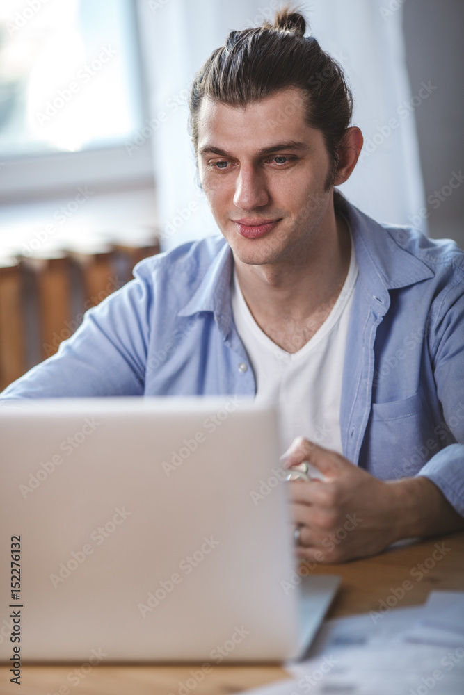 Young man holding mug, sitting at the table and looking to the laptop screen
