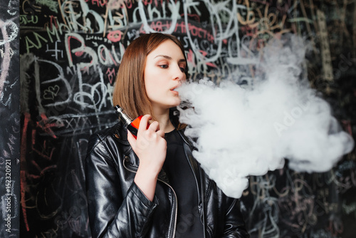 Beautiful young woman inhaling smoke. young girl vaping against the background of a painted wall