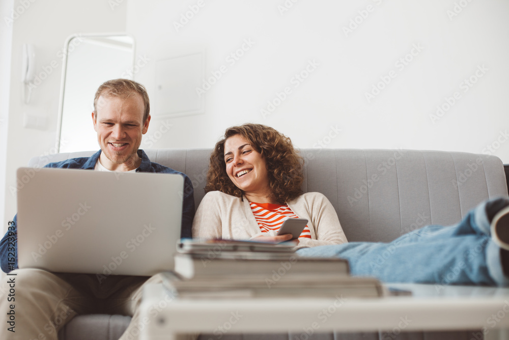 Family spending time with electronic devices. Man using laptop and smiling woman typing message with mobile phone. Sitting together on the sofa Family spending time with electronic devices. Man using laptop and smiling woman typing message with mobile phone. Sitting together on the sofa