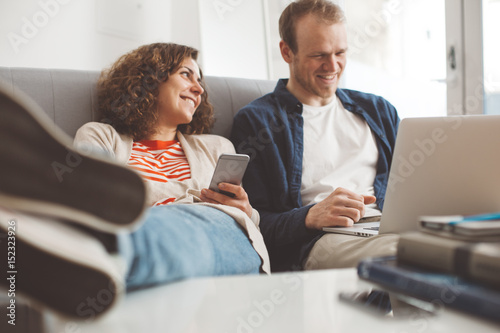 Happy family spending time with electronic devices. Man using laptop and smiling woman typing message with mobile phone. Sitting together on the sofa