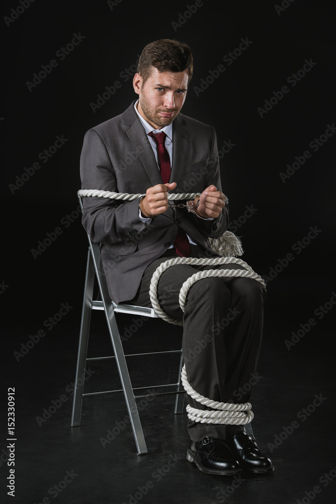 young upset businessman roped on to chair isolated on black young upset businessman roped on to chair isolated on black