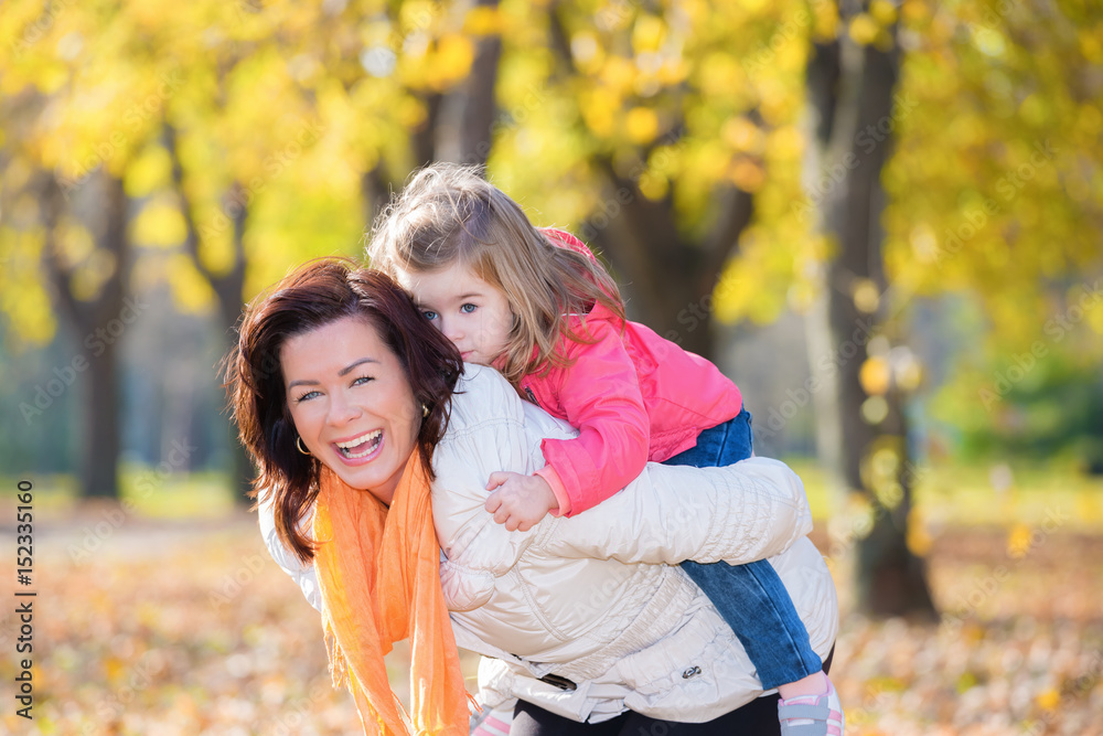 Mother giving piggy back to her little daughter in the autumn park Mother giving piggy back to her little daughter in the autumn park