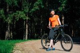 Young pretty woman in helmet and orange shirt with bicycle resting and sunbathing on the forest road after ride. Healthy lifestyle concept. Sports Girl with bike.