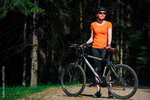 Young pretty woman in helmet and sunglasses standing with bicycle on the forest road. Healthy lifestyle concept. Girl with bike.