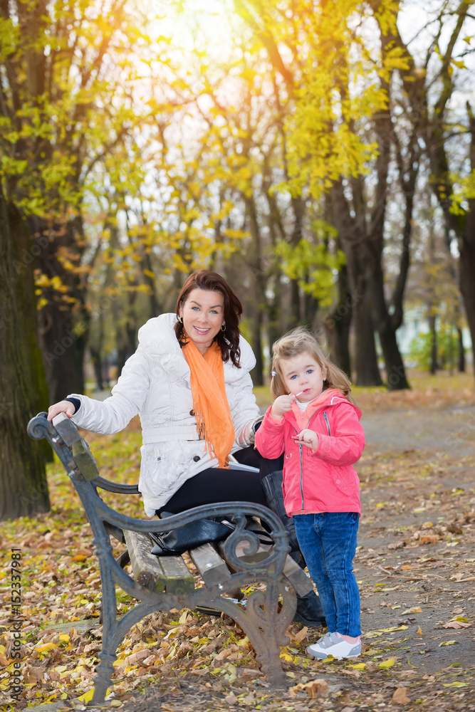 Mother and daughter resting in the autumn park.Little girl applying lips balm.Focus on the woman face