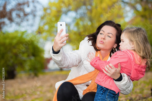 Mother and little daughter taking funny selfie in autumn park.Focus on the child