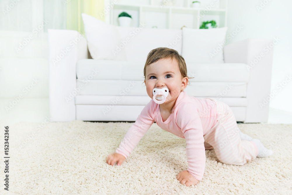 Happy baby little girl crawling with a pacifier in a mouth on the living room floor, looking at camera.Copy space Happy baby little girl crawling with a pacifier in a mouth on the living room floor, looking at camera.Copy space
