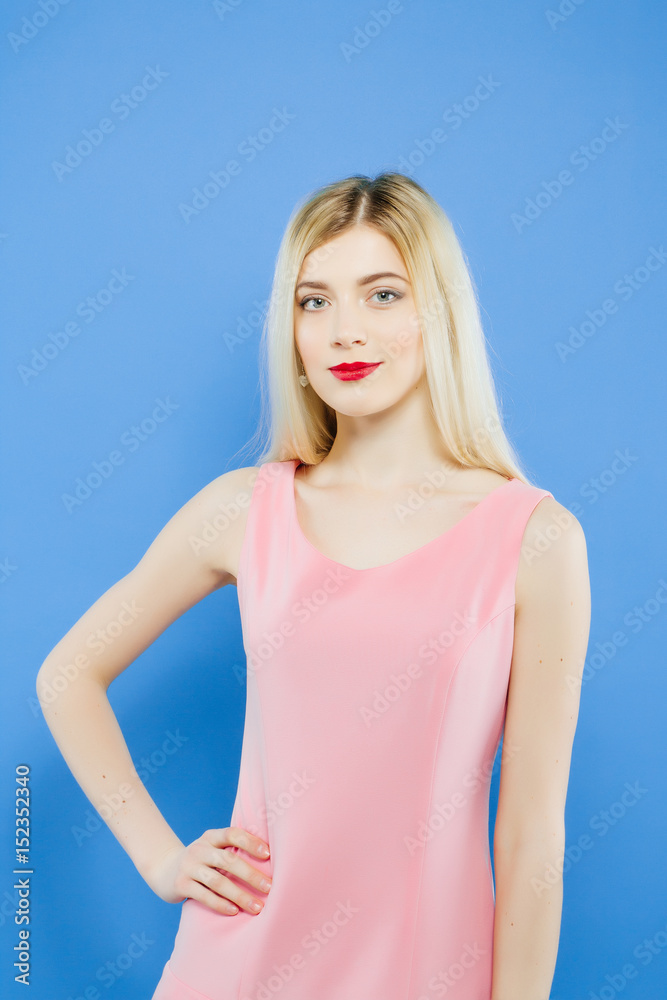 Beautiful Young Woman with Charming Smile in Pink Dress is Posing in Studio. Portrait of Pretty Blonde on Blue Background. Beautiful Young Woman with Charming Smile in Pink Dress is Posing in Studio. Portrait of Pretty Blonde on Blue Background.