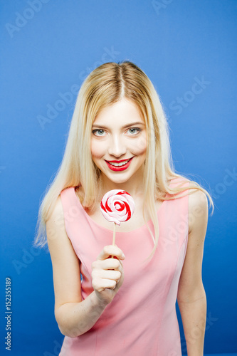 Playful Girl Wearing Pink Dress Wants to Bite a Peace of Lollipop. Cute Blonde is Posing in Studio on Blue Background. © Andriy