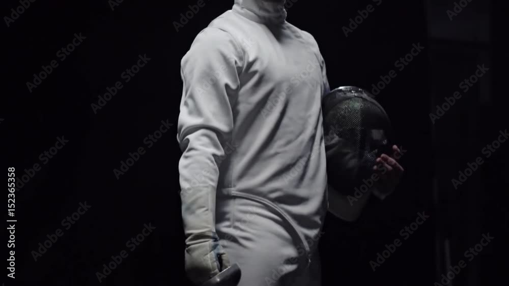 Tilt up of professional fencer with protective mask and foil standing and looking at camera Tilt up of professional fencer with protective mask and foil standing and looking at camera