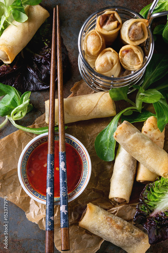 Fried spring rolls with red pepper sauce, served on crumpled paper and in fry basket with fresh green salad and wooden chopsticks over old metal texture background. Top view. Asian food