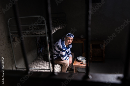 View through iron door with prison bars on male wearing prison uniform sitting on a bed