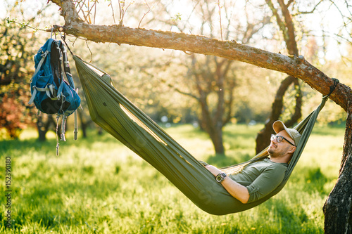 Happy traveler bearded hipster man in green hat and glasses lying in hummock on apple tree in blooming garden in summer sunny day on vacation. Weekend. Tourism. Leisure. Dreaming. Fragrance. Holiday.