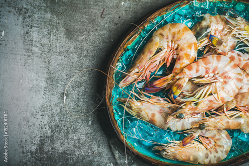 Raw uncooked tiger prawns on chipped ice in turquoise blue ceramic tray over grey concrete background, top view, copy space, horizontal composition. Fresh seafood