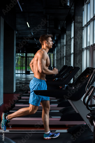 Side view full length of young man in sportswear running on treadmill at gym