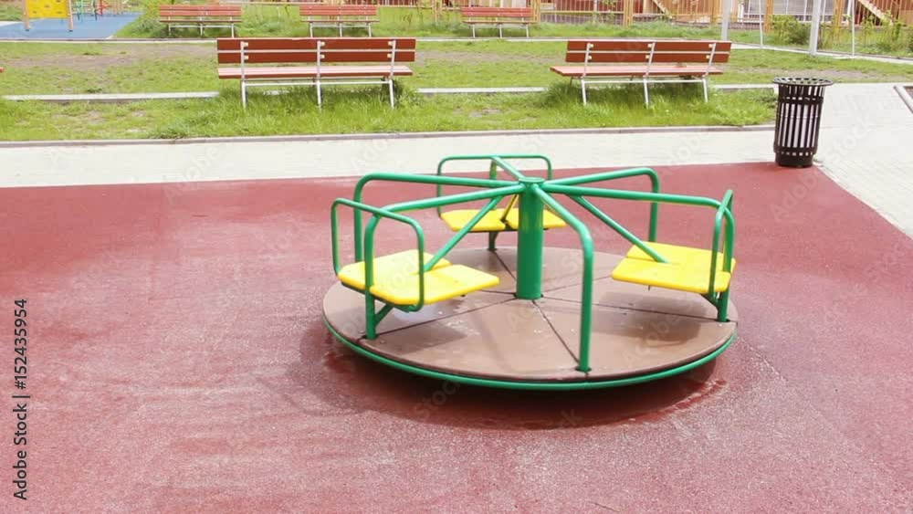 Yellow with green color, a wet carousel is spinning alone on a rubberized burgundy playground. no people. In the background high residential buildings of high-rise buildings Yellow with green color, a wet carousel is spinning alone on a rubberized burgundy playground. no people. In the background high residential buildings of high-rise buildings