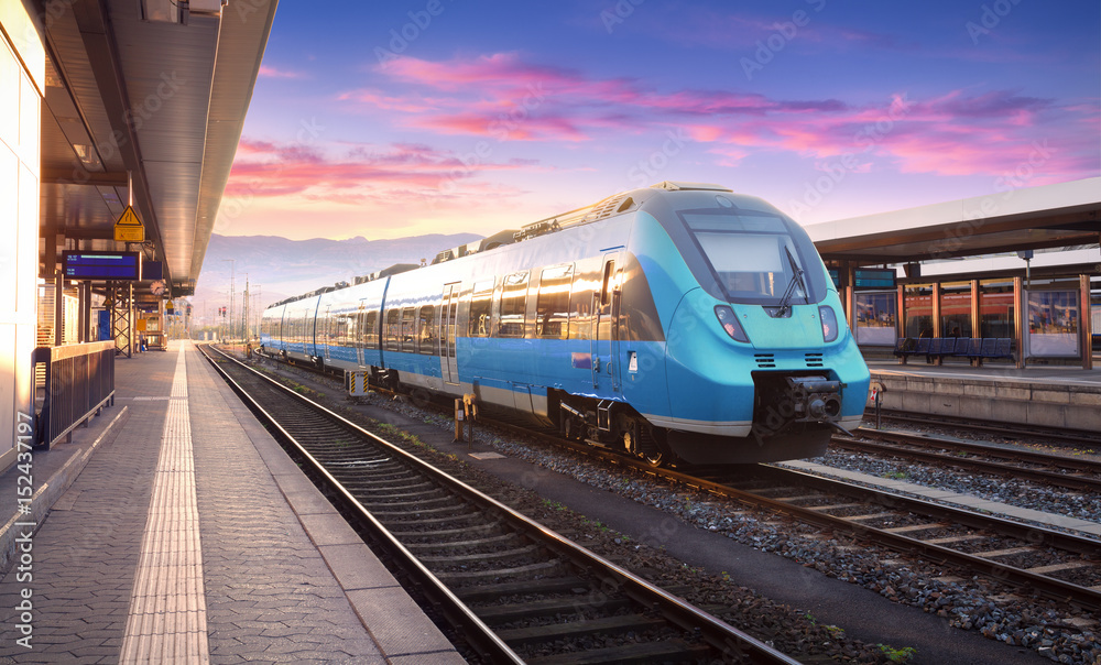 Obraz premium Piękny widok z nowoczesnym pociągiem dużych prędkości na stacji kolejowej i kolorowe niebo z chmurami o zachodzie słońca w Europie. Przemysłowy krajobraz z błękita pociągiem na kolejowej platformie. Tło kolejowe