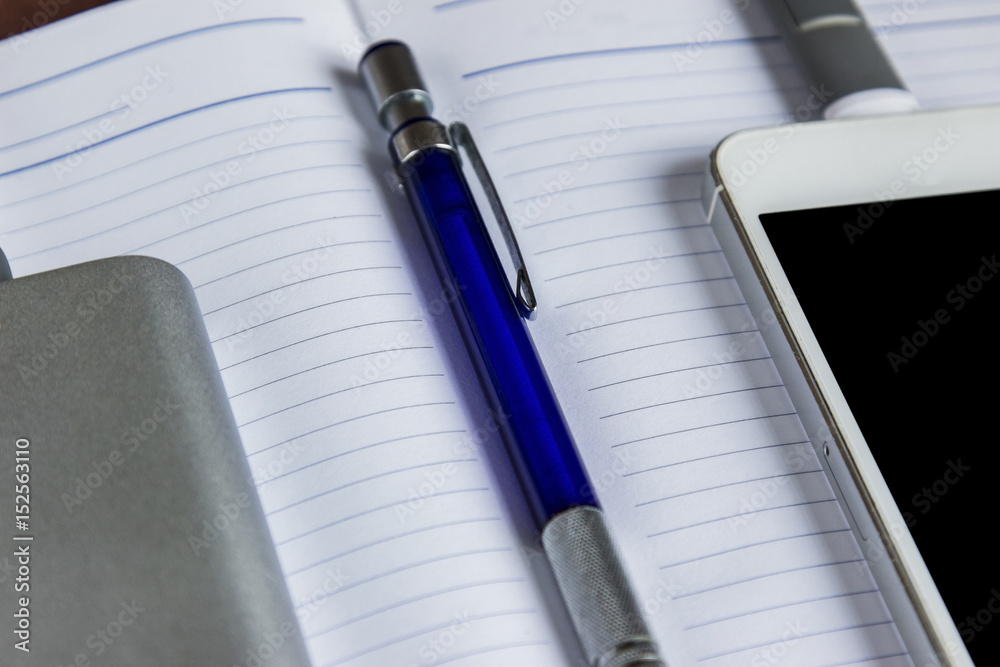 Charging Smartphone With Grey Portable External Battery And Pen On A Open Notebook Charging Smartphone With Grey Portable External Battery And Pen On A Open Notebook
