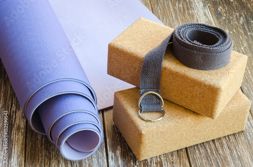 Accessories or props for yoga, pilates or fitness. Exercise lilac mat, two cork blocks and grey strap on wooden background.