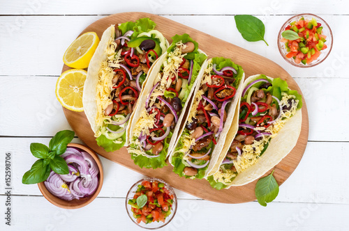 Tacos is a traditional Mexican dish. Tortilla stuffed with chicken, bell and hot peppers, beans, lettuce, cheese, blue onion with salsa sauce on white wooden background. Top view.